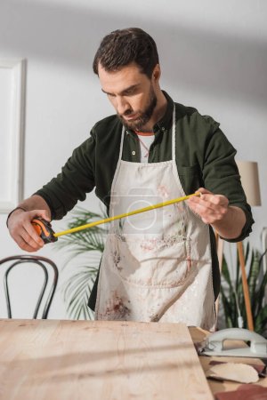 Téléchargez les photos : Restaurateur barbu dans un tablier sale regardant la règle près de la planche en bois dans l'atelier - en image libre de droit