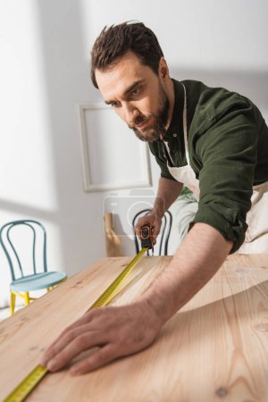 Bearded carpenter in apron measuring wooden board in workshop 