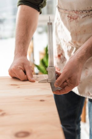 Téléchargez les photos : Vue recadrée de l'artisan dans un tablier sale mesurant la planche en bois avec étriers dans l'atelier - en image libre de droit