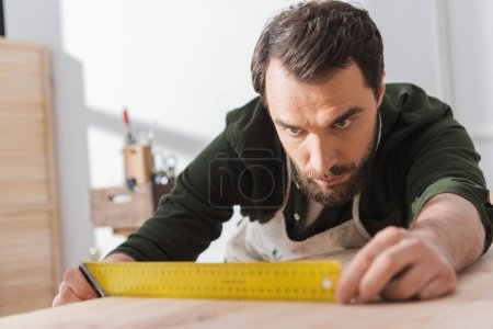 Foto de Focused carpenter looking at square tool on blurred wooden board - Imagen libre de derechos