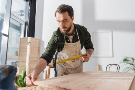 Téléchargez les photos : Carpenter in apron holding square tool and touching wooden board in workshop - en image libre de droit