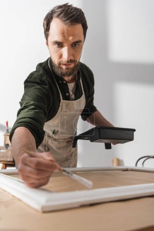 Foto de Focused restorer in apron holding paintbrush near blurred picture frame in workshop - Imagen libre de derechos