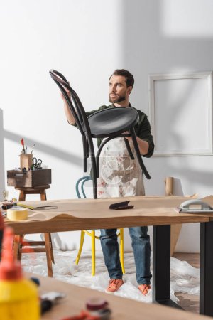 Foto de Bearded restorer holding chair near working table in workshop - Imagen libre de derechos