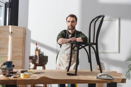 Bearded restorer in apron looking at camera near wooden chair in workshop 