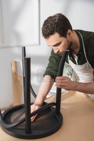 Handwerker in Schürze poliert schwarzen Holzstuhl in Werkstatt 