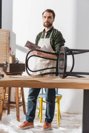 Foto de Restaurador sosteniendo papel de lija y mirando la cámara cerca de la silla de madera en el taller - Imagen libre de derechos