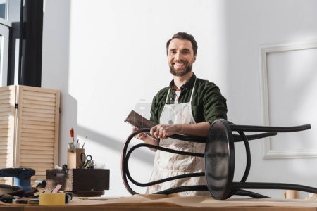 Photo for Smiling restorer in apron holding sandpaper near chair and looking at camera in workshop - Royalty Free Image