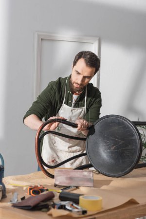 Bearded restorer sanding chair near blurred tools on table in workshop 