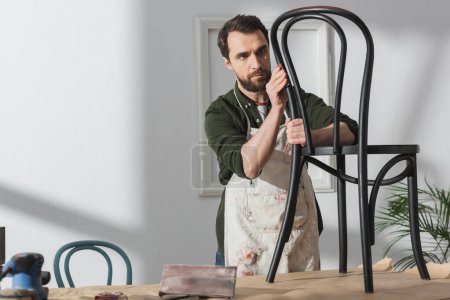 Bearded craftsman in apron looking at wooden chair near sandpaper and sanding machine in workshop 