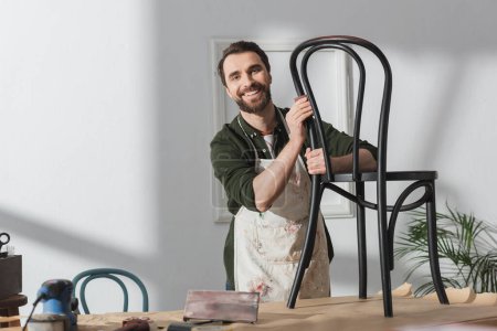 Cheerful restorer in apron looking at camera near chair and sandpaper in workshop 