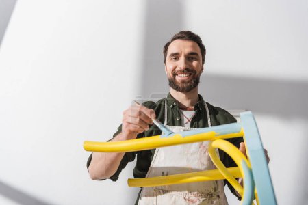 Smiling bearded restorer looking at camera while repainting chair in workshop 