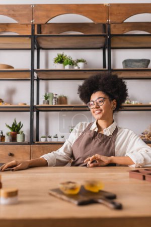 joyful african american craftswoman in apron and eyeglasses sitting near blurred handmade soap on table in workshop