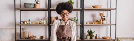 mujer afroamericana feliz en gafas y delantal de pie cerca de estante borroso con libros e ingredientes naturales en taller de artesanía, pancarta
