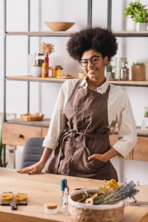 happy african american woman with hand in pocket of apron standing near table with blurred soap bars and dried herbs 