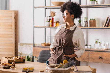 Téléchargez les photos : Femme afro-américaine avec les mains dans des poches de tablier debout près de savon fait à la main et des ingrédients dans l'atelier d'artisanat - en image libre de droit