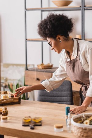 Téléchargez les photos : Femme afro-américaine dans les lunettes et tablier atteignant savonnettes près des ingrédients flous sur la table dans l'atelier d'artisanat - en image libre de droit
