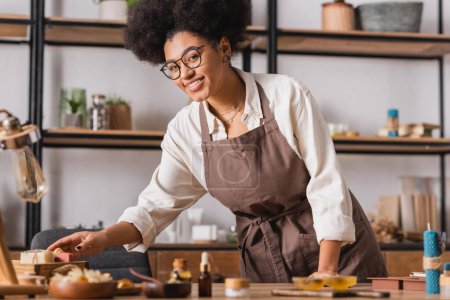 souriant artisan afro-américain dans des lunettes et tablier atteignant savon fait main près des ingrédients flous sur la table