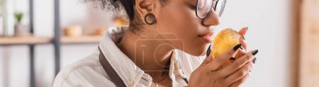 geschnittene Ansicht einer afrikanisch-amerikanischen Kunsthandwerkerin in Brille, die den Geschmack handgemachter Seife in der Werkstatt genießt, Banner