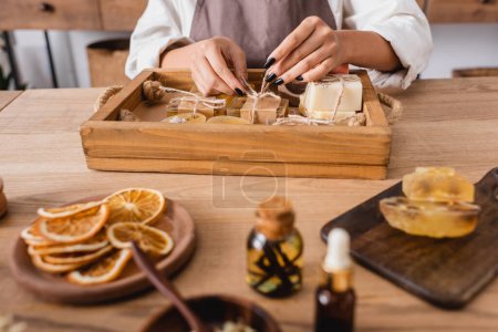 vista recortada de la artesana afroamericana atando cordel en jabón hecho a mano cerca de rebanadas de naranja seca y aceites esenciales en primer plano borroso