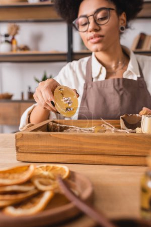 Foto de Blurred african american woman in eyeglasses holding handmade soap near wooden tray in craft workshop - Imagen libre de derechos