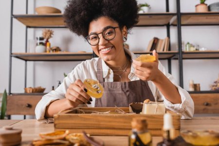 happy african american craftswoman in eyeglasses holding handmade soap near natural ingredients on blurred foreground