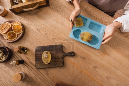 top view of cropped african american woman holding silicone mold with handmade soap near chopping board and natural ingredients