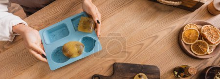 Téléchargez les photos : Vue du dessus de la femme afro-américaine cultivée tenant un moule en silicone avec des barres de savon près de tranches d'orange séchées sur une table en bois, bannière - en image libre de droit