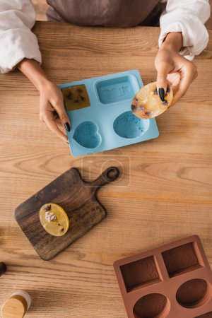 top view of cropped african american woman holding handmade soap near silicon molds and cutting board on wooden table