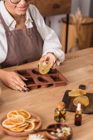 Foto de Partial view of african american craftswoman with handmade soap near silicone mold and natural ingredients on wooden table - Imagen libre de derechos
