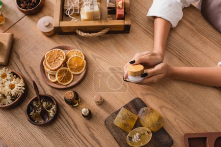 Téléchargez les photos : High angle view of cropped african american woman holding cosmetic cream near handmade soap and natural ingredients on wooden table - en image libre de droit