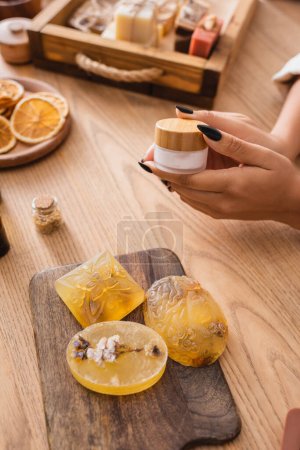 partial view of african american woman holding cosmetic cream near handmade soap on wooden chopping board