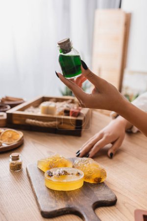 cropped view of african american woman holding jar with essential oil near handmade soap on chopping board