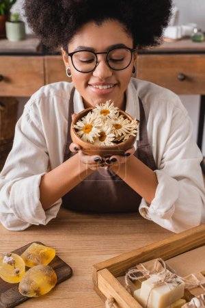 happy african american craftswoman in eyeglasses holding wooden bowl with dried camomiles near bars of handmade soap