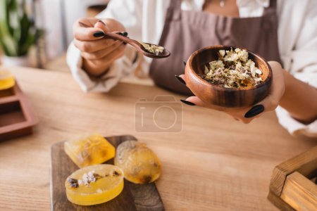 cropped view of african american craftswoman holding wooden bowl and spoon with dried plants near herbal homemade soap
