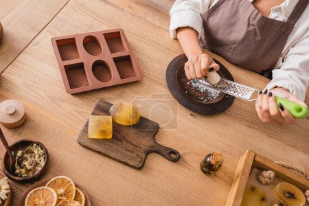 vista superior del jabón de rejilla de mujer afroamericana recortado cerca del molde de silicona y los ingredientes naturales en la mesa de madera