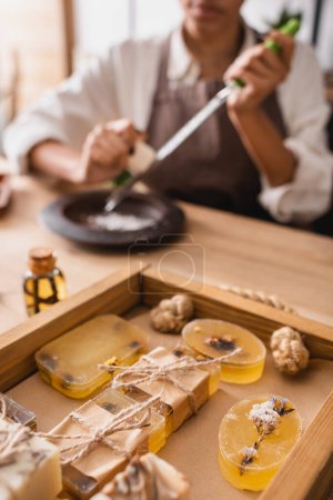 Foto de Selective focus of wooden tray with herbal soap near african american craftswoman on blurred background - Imagen libre de derechos