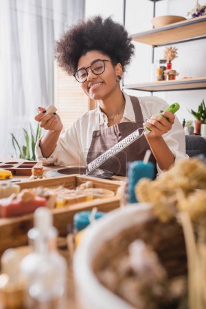 cheerful african american woman with soap bar and grater looking at camera on blurred foreground in crafts workshop