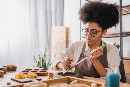 Téléchargez les photos : Young african american craftswoman in eyeglasses and apron grating soap near silicone mold in workshop - en image libre de droit