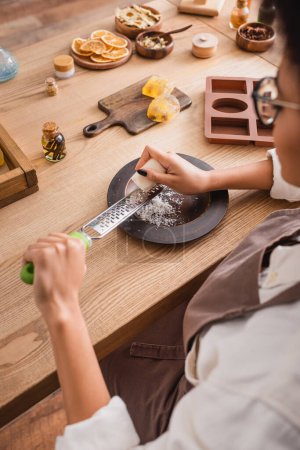 vista recortada de borrosa africana americana artesana rejilla jabón cerca de molde de silicona e ingredientes naturales en mesa de madera