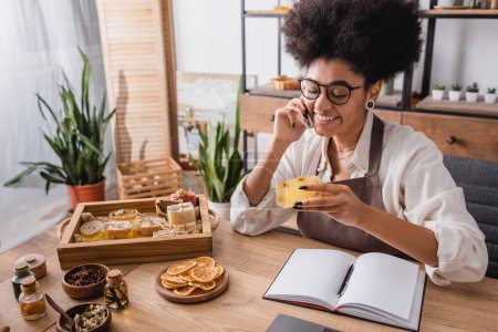 heureuse femme afro-américaine tenant du savon fait maison et parlant sur téléphone portable près d'ingrédients naturels et ordinateur portable