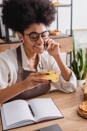 Téléchargez les photos : Cheerful african american craftswoman in eyeglasses holding handmade soap while talking on smartphone near notebook - en image libre de droit