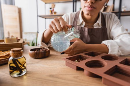 vista parcial de la mujer afroamericana sosteniendo jarra con jabón líquido cerca de molde de silicona e ingredientes naturales en taller de artesanía