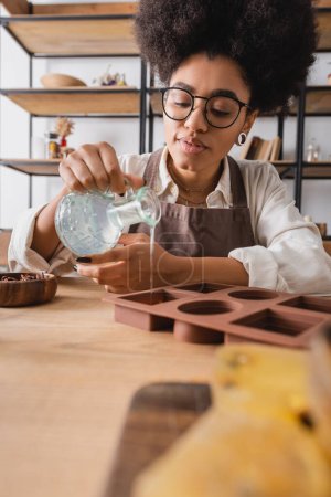 Afrikanisch-amerikanische Kunsthandwerkerin in Brille gießt Flüssigseife in Silikonform auf verschwommenem Vordergrund in Werkstatt