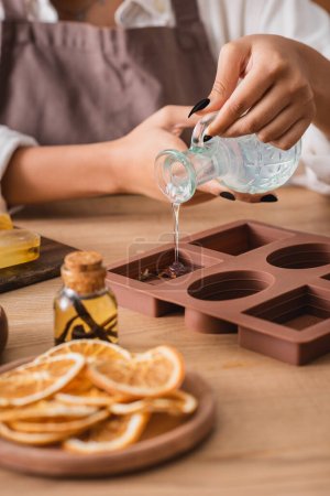 cropped view of african american craftswoman pouring liquid soap in silicone mold near dried orange slices and essential oil