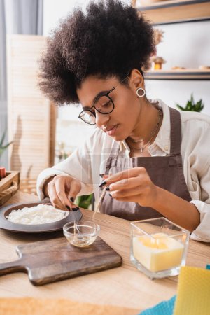 african american woman in eyeglasses dipping twine in melted wax near chopping board and burning candle