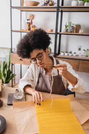 african american craftswoman in apron and eyeglasses holding candle wick near natural wax sheet and parchment on wooden table magic mug #640941116