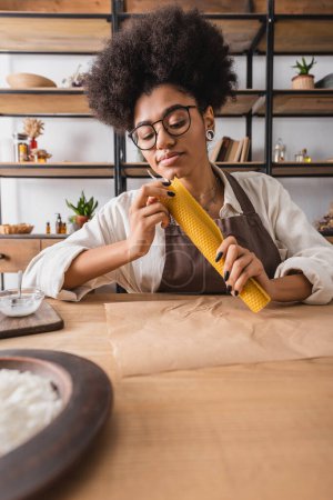 artesana afroamericana rizada en delantal y anteojos haciendo mango natural cerca de pergamino en mesa en taller
