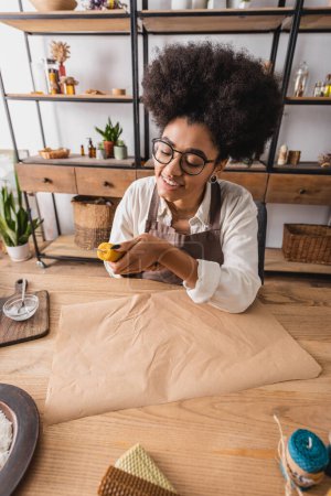 lächelnde afrikanisch-amerikanische Kunsthandwerkerin in Schürze und Brille, die in der Werkstatt Naturkerzen in der Nähe von Bastelpapier herstellt