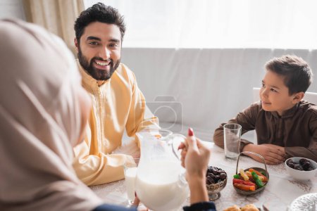 Photo for Smiling muslim father and son looking at blurred woman in hijab during suhur at home - Royalty Free Image