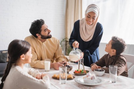 Foto de African american woman in hijab pouring milk near family during suhur breakfast at home - Imagen libre de derechos